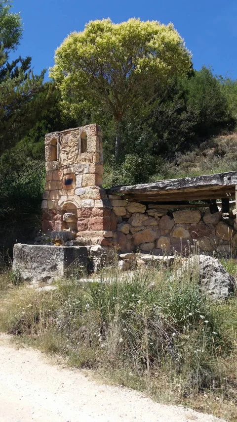Fuente rústica de piedra dedicada a Félix Rodríguez de la Fuente junto a cobertizo y árbol
