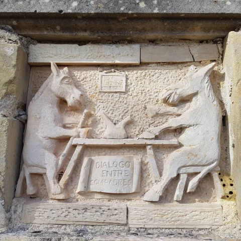 Relieve de piedra tallado con dos burros sentados a una mesa titulado Diálogo entre compadres
