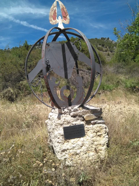Escultura esférica de metal con corazón y alas sobre pedestal de piedra en campo