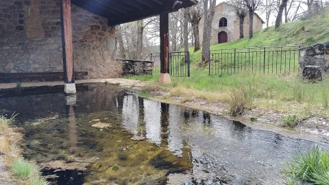 Pozal de agua del lavadero techado con pilares de madera y la ermita de San Miguel al fondo en la ladera