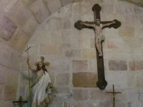 Crucifijo de madera adosado a la pared de piedra junto a escultura de Cristo Resucitado con estandarte