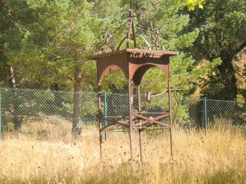 Estructura de hierro forjado sobre pozo con detalles decorativos calados en el prado de la ermita en Mamolar