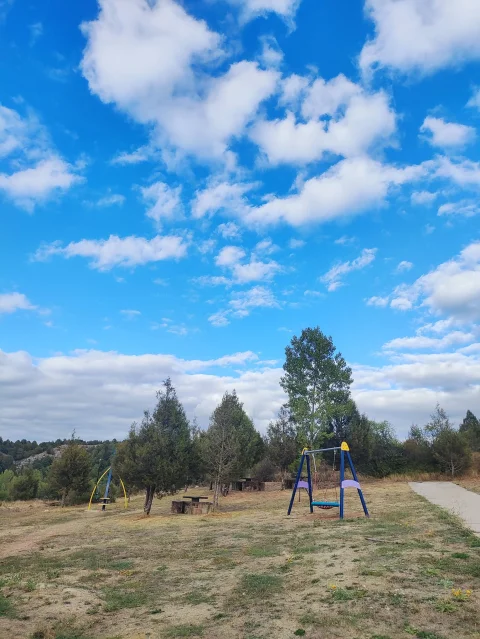 Columpios infantiles y mesas de pícnic en pradera arbolada bajo cielo azul con nubes en Mamolar