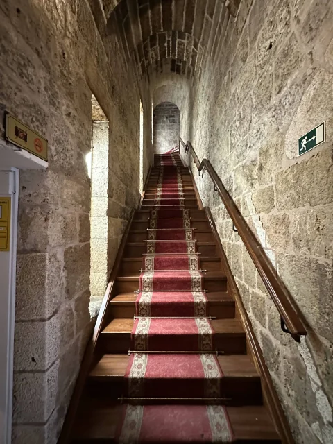 Escalera de madera con alfombra roja bajo bóveda de cañón apuntada en sillería de piedra