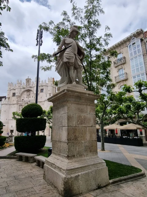 Vista angular de la estatua de Fernando I sobre pedestal de sillería con el Arco de Santa María de fondo