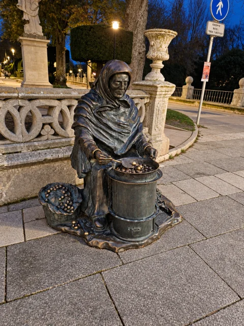 Estatua de bronce de la castañera de noche sentada junto a su hornillo bajo la luz de una farola urbana