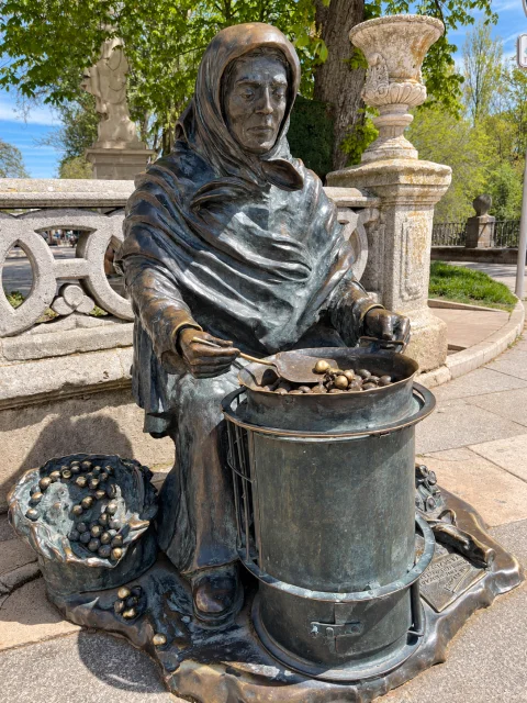 Plano medio de escultura de bronce de castañera removiendo castañas con paleta frente a balaustrada