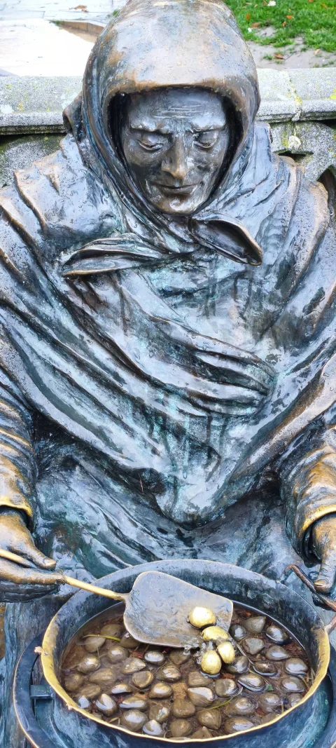 Primer plano de estatua de bronce con detalle del rostro arrugado de anciana y sartén con castañas asadas