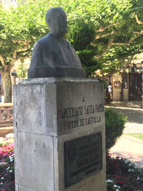 Busto de bronce de perfil sobre pedestal con inscripción conmemorativa entre flores y ramas verdes frondosas