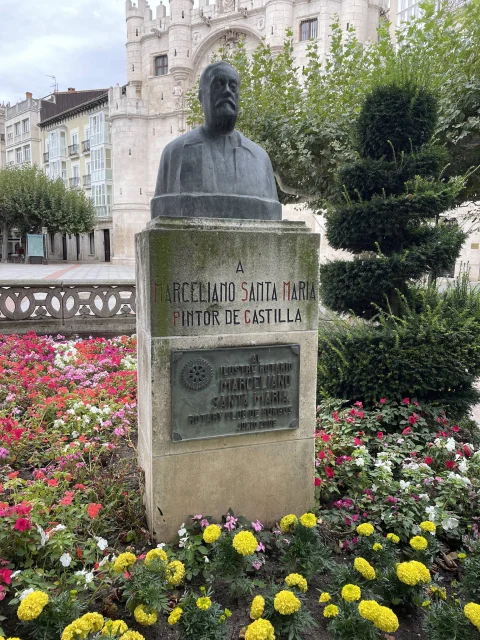 Busto de Marceliano Santa María enmarcado por parterres floridos y ciprés esculpido ante el Arco