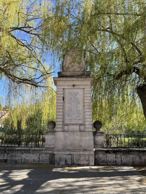 Monolito neoclásico con lápida conmemorativa en latín y escudo episcopal bajo las ramas de sauce