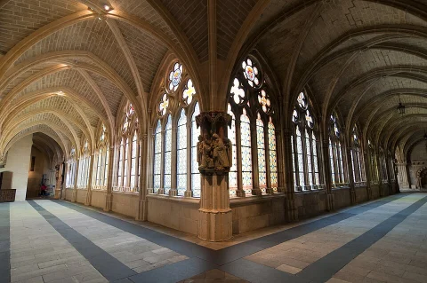 Intersección de galerías góticas del claustro alto con suelo de bandas oscuras, pilar historiado y vidrieras