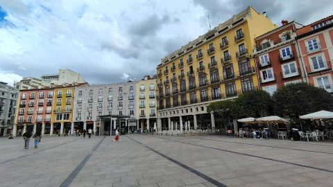 Plaza urbana amplia con soportales, terrazas de cafetería bajo sombrillas y fachada alta de color amarillo