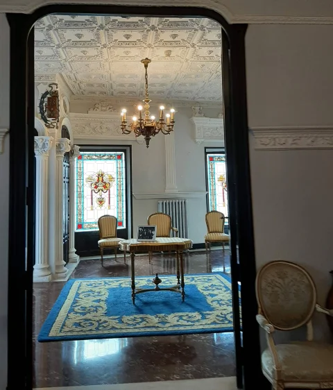 Encuadre desde el pasillo hacia una elegante sala con lámpara de araña, sillas de época, mesa auxiliar y vidrieras