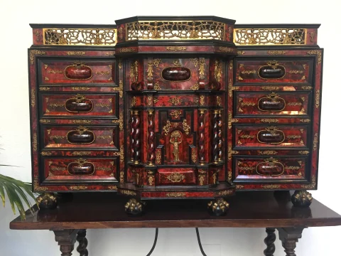 Plano frontal de un mueble bargueño con múltiples cajones decorados en rojo y bronce, patas de garra y barandilla superior de latón