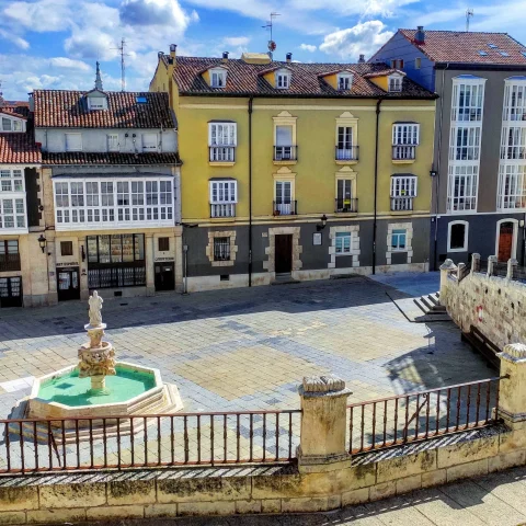 Barandilla de piedra en primer plano sobre la plaza con fuente octogonal y casas de colores en Burgos