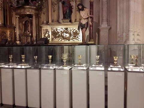 Hilera de vitrinas individuales con cálices litúrgicos de plata dorada ante un retablo barroco con Cristo atado a la columna