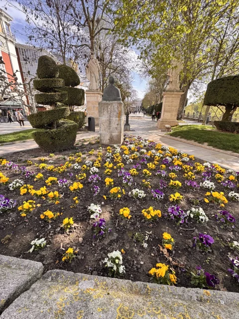 Cama de flores violetas y amarillas con busto de bronce sobre pedestal y estatuas de reyes al fondo