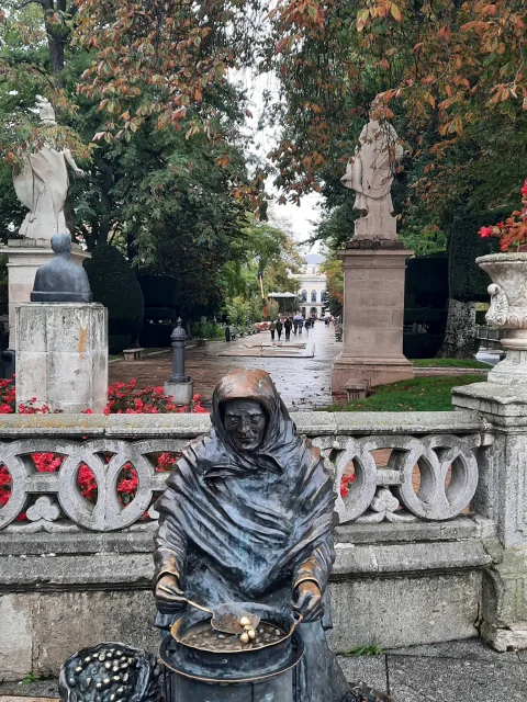 Escultura de bronce de la Castañera asando castañas sobre la barandilla con las estatuas al fondo