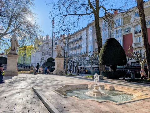 Chorro de agua saliendo de una fuente baja de piedra caliza con estatuas y edificios blancos al fondo