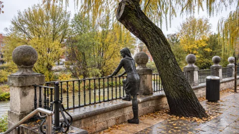 Estatua en bronce de «Mujer mirando al río Arlanzón» apoyada en el pretil bajo un sauce llorón otoñal