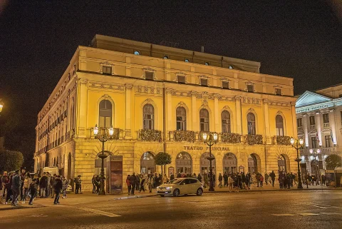 Fachada de teatro clásico iluminada de amarillo por la noche con viandantes y vehículos en la plaza