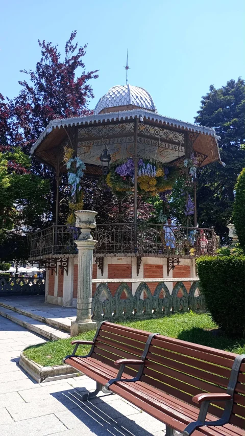 Banco de madera en primer plano ante el templete de música de hierro decorado con flores moradas