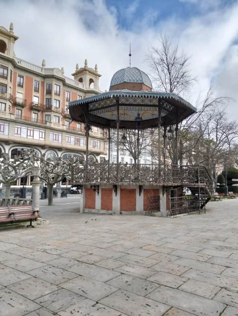 Quiosco de música de hierro con barandilla calada y escalinata en una plaza de piedra ante edificios