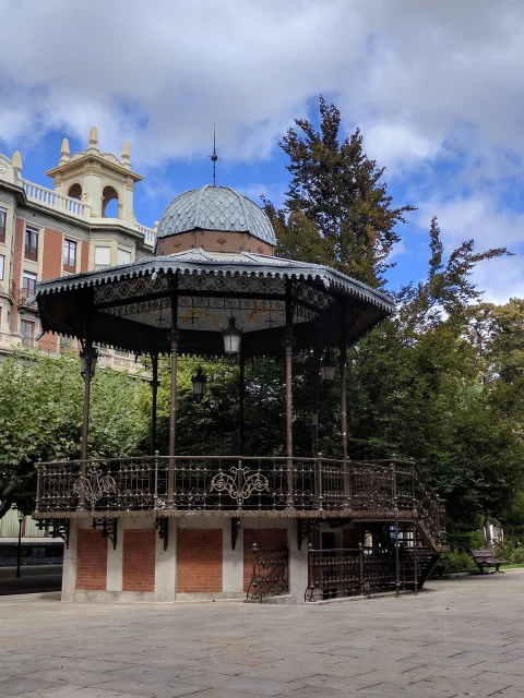 Vista en contrapicado del quiosco de música de forja con base de ladrillo y edificios clásicos al fondo