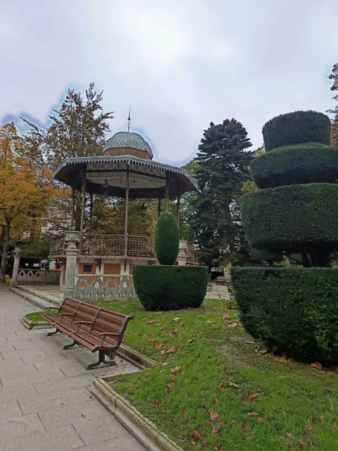 Vista lateral del quiosco de música ochavado entre bancos de madera y cipreses esculpidos