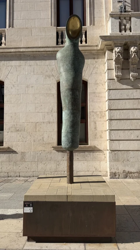 Vista frontal de la escultura estilizada con hueco dorado sobre pedestal cúbico de piedra y acero corten