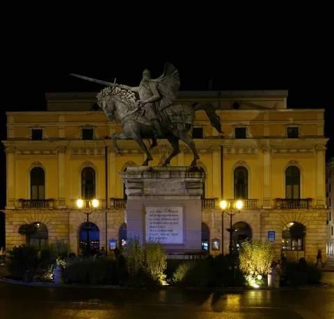 Monumento ecuestre de bronce iluminado de noche en una rotonda frente a la fachada de un teatro clásico