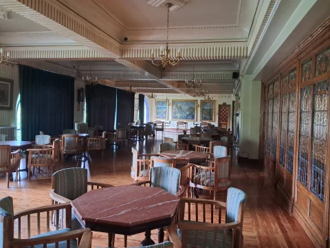 Amplio salón social con mesas octogonales de mármol rojizo, butacas de madera tapizadas y cuadros al óleo