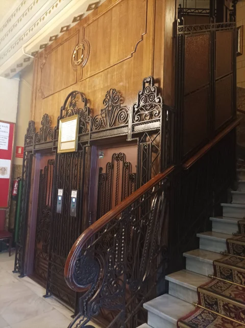 Antigua cabina de ascensor con artesonado de madera de nogal, cerrajería modernista de forja y escalera alfombrada