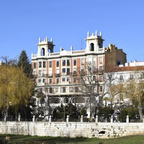 Edificio señorial de ladrillo rojo y torres blancas visto detrás de árboles y muro de piedra sobre río