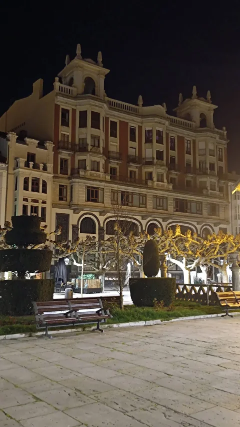 Fachada regionalista iluminada de noche con setos topiarios y bancos del paseo peatonal