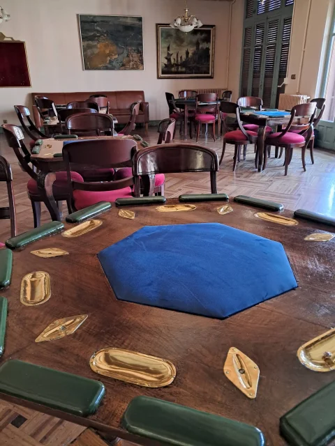 Mesa octogonal de madera con tapete azul en primer plano y salón con mesas y sillas de terciopelo