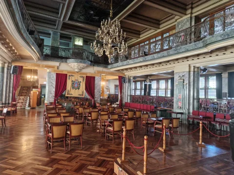 Salón señorial con mesas de café, barandilla de forja en el piso superior y gran lámpara dorada colgada