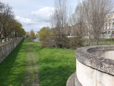 Pretil curvo de piedra en primer plano con vistas a pradera verde, álamos y cauce de un río