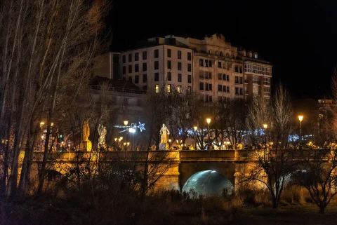 Puente iluminado de noche con alumbrado navideño, estatuas del ciclo cidiano y fachadas históricas