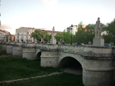 Tajamares y arcos de piedra con estatuas cidianas y edificios señoriales al fondo durante el atardecer