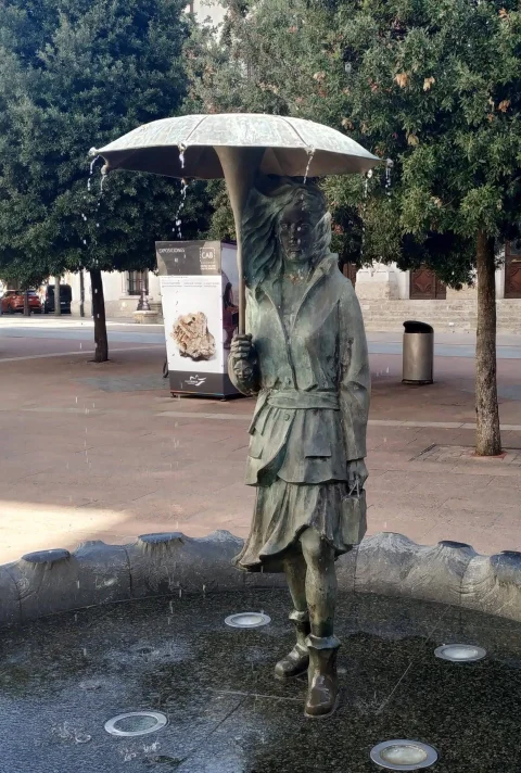 La mujer de bronce con gabardina y botas bajo el paraguas abierto, de frente, en el centro del pilón