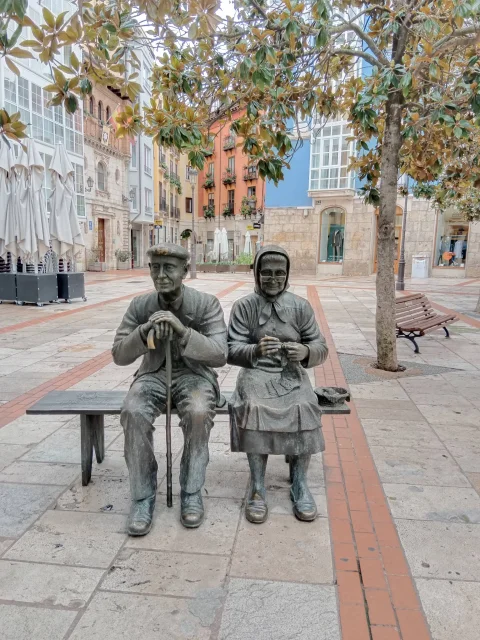 Los Abuelos sentados de Burgos bajo la magnolia, con las fachadas de colores de la calle al fondo