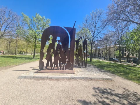 Escultura de acero corten oxidado sobre base de adoquines en un parque arbolado