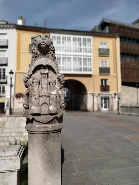Primer plano de un poste de piedra esculpido con un león y un castillo en relieve, con el Arco de San Juan