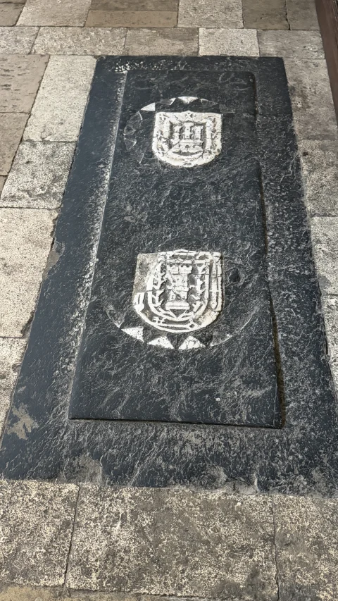 Losa funeraria de piedra oscura encastrada en el pavimento con dos escudos heráldicos labrados en bajo relieve