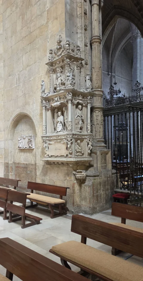 Rincón de la capilla con bancos de madera, pila en primer plano y templete o monumento de piedra adosado a la columna