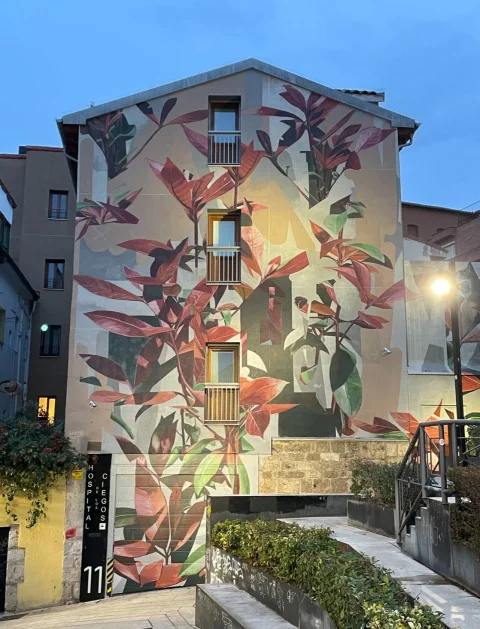 Vista frontal del edificio residencial con mural de hojas rojizas y verdes iluminado por farolas urbanas al anochecer