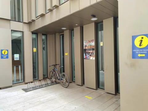 Acceso peatonal a la oficina de turismo con señalización amarilla, aparcabicicletas metálico