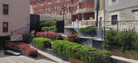 Zona ajardinada con rampas escalonadas, arbustos verdes y rojizos, barandillas metálicas